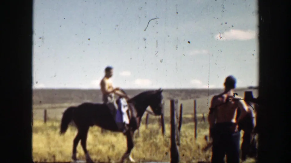 Two shirtless men on horses pause or ride near fence posts in a dry rural California field, filmed in soft 8mm color.