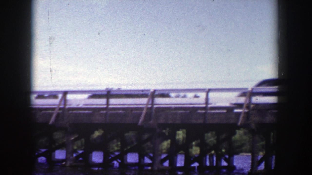 Car crossing a wooden bridge above water, framed by dark 8mm film borders.