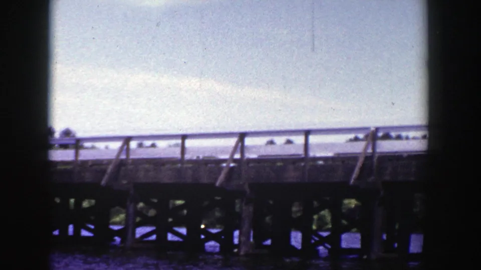 1953 Wisconsin home-movie frame of a car crossing a wooden bridge over water, partly hidden by a dark vertical frame edge.