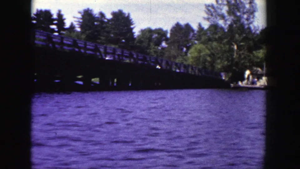 1953 Wisconsin 8mm frame showing the wooden bridge over rippled water with trees and shoreline beyond.