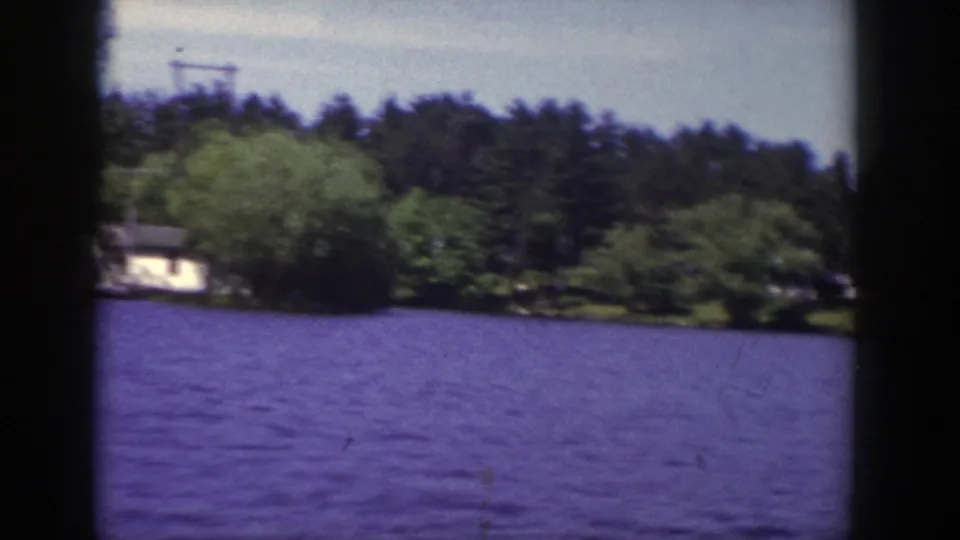 1953 Wisconsin 8mm frame showing a wooded lakeshore and small house across rippled lake water.