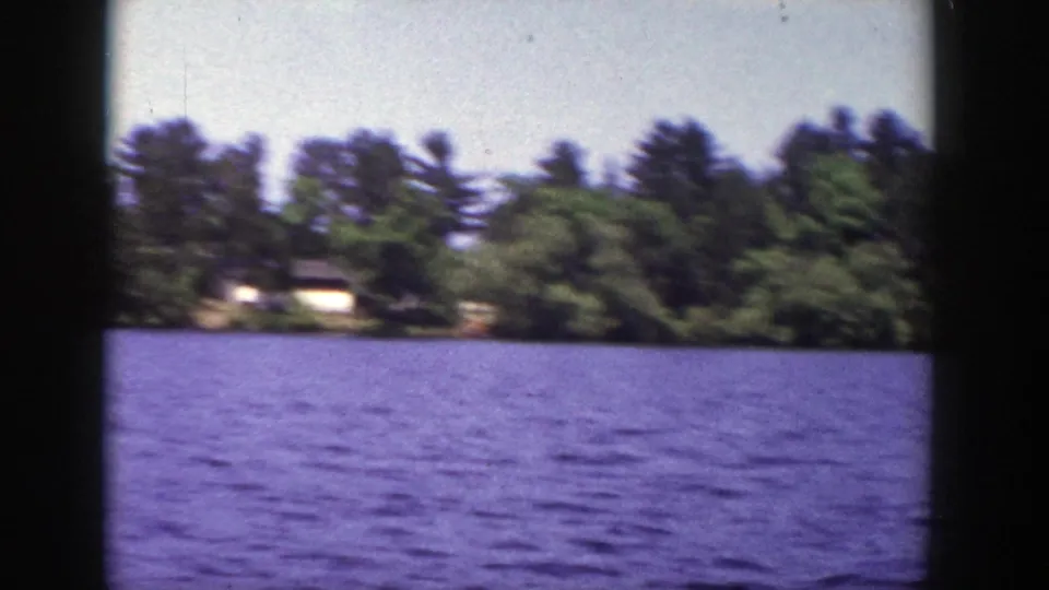1953 Wisconsin 8mm frame of a quiet lake surface and wooded shoreline with a small distant house.
