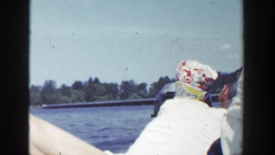 1953 Wisconsin boating view with a seated passenger in a patterned headscarf facing the lake and distant tree-lined shore.