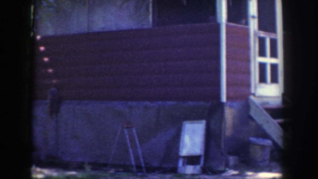 1953 Wisconsin 8mm frame showing a red-sided house exterior with a white door, small steps, ladder, and yard objects.