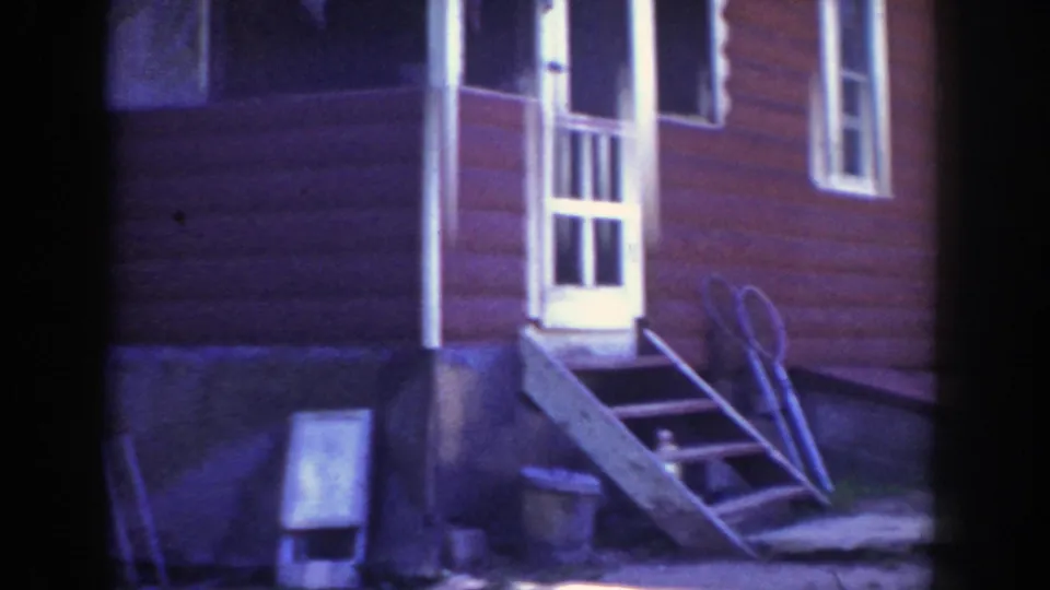 1953 Wisconsin home-movie frame of a red house side with white doorway, steps, ladder, and objects against the foundation.