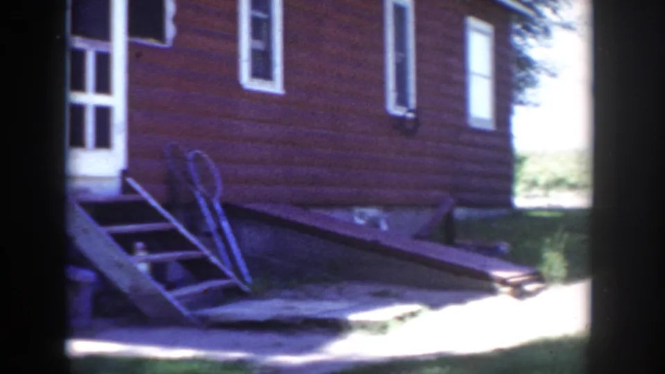 1953 Wisconsin 8mm frame showing the side of a red house, white-trimmed openings, a tipped ladder, walkway, and sunlit grass.