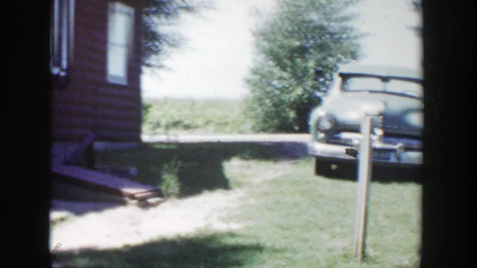 1953 Wisconsin home-movie frame with red house siding, grassy yard, tree, clothesline post, and an older vehicle parked beside the house.