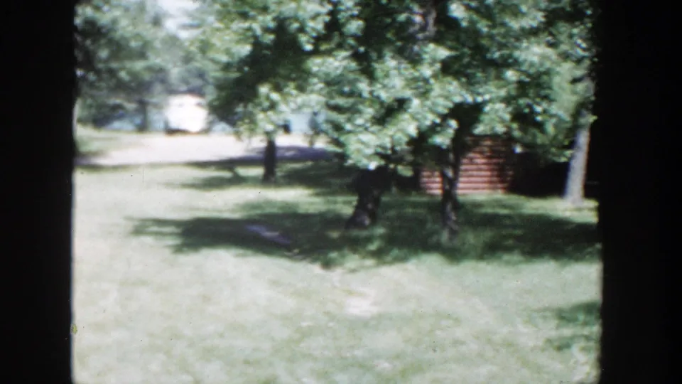 Late frame of the 1953 Wisconsin backyard pan, showing shaded grass, leafy trees, a distant pale car, and a structure at right.