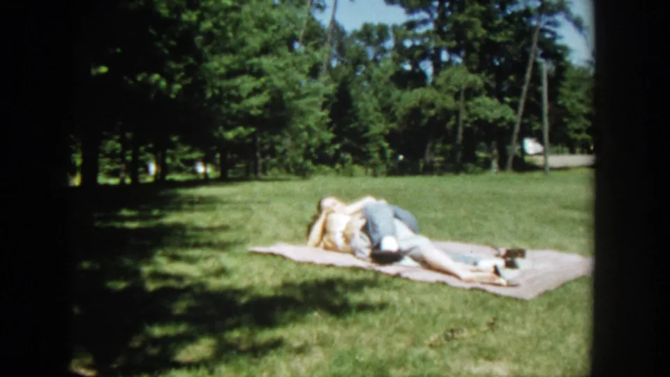 A 1953 Wisconsin home-movie frame shows the couple lying close on a blanket, partly shaded by trees in a park.