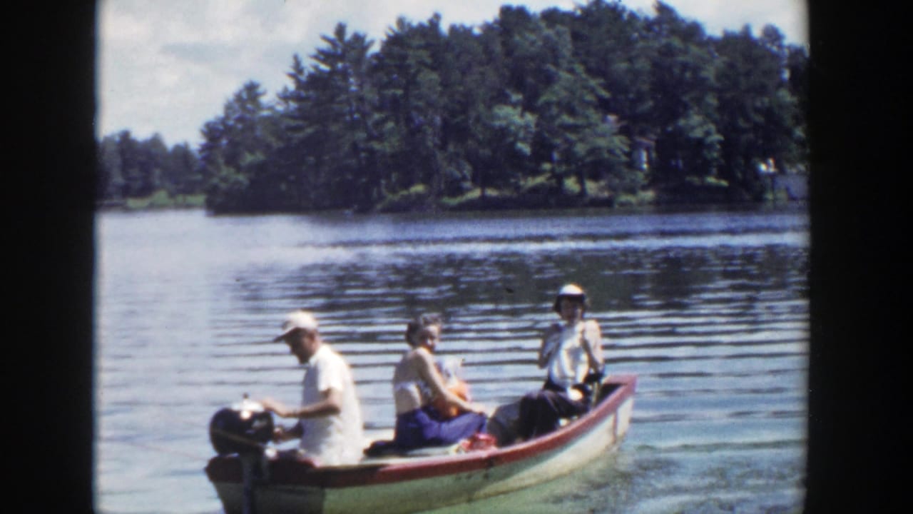 In 1953 Wisconsin home-movie footage, three adults sit in a small outboard motorboat moving across a calm lake.