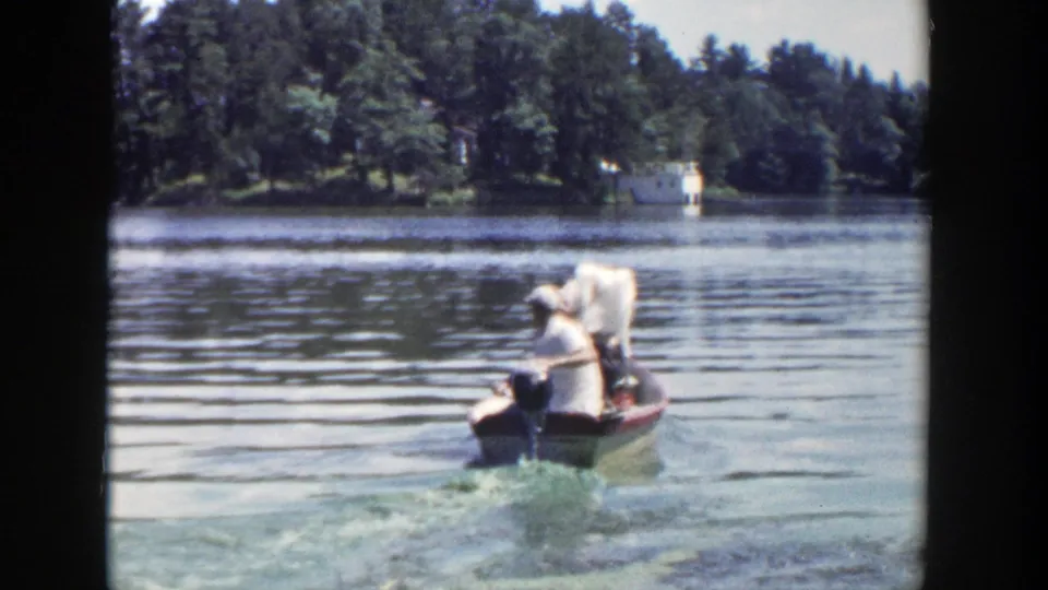 The boat recedes across the Wisconsin lake, with three adults aboard and a wooded shoreline beyond the wake.