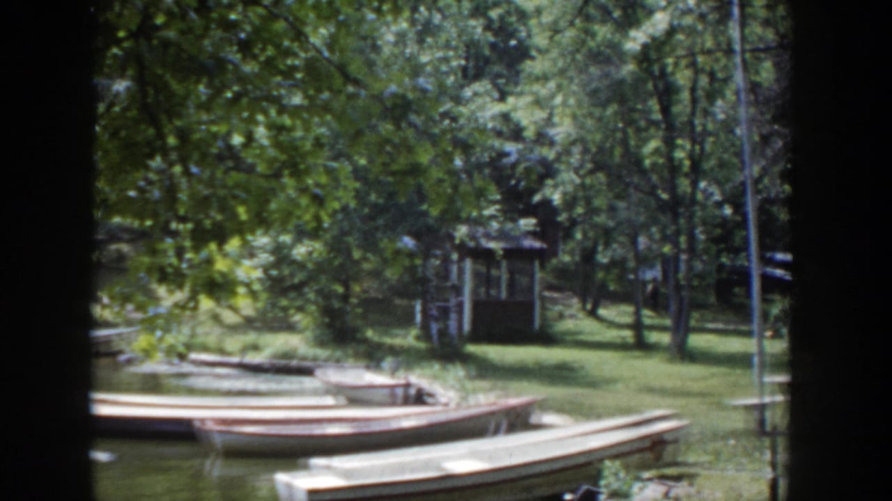 1953 Wisconsin 8mm frame of a shaded lakeside park, with wooden rowboats by the shore and a small rustic structure beyond the trees.