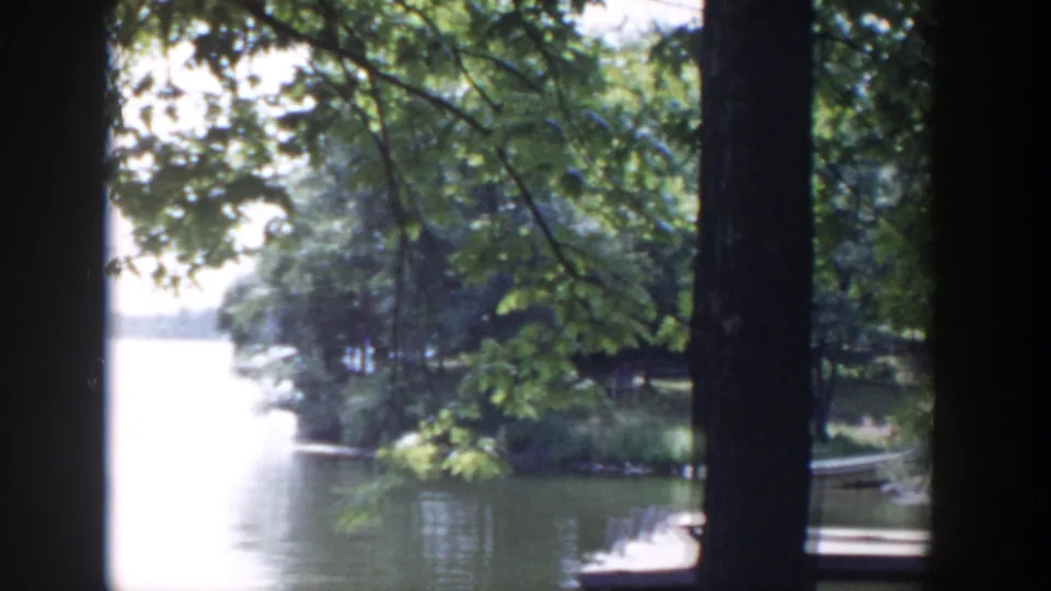 1953 Wisconsin 8mm frame showing the lake opening through shaded trees, with shoreline vegetation and rowboats near a dock.