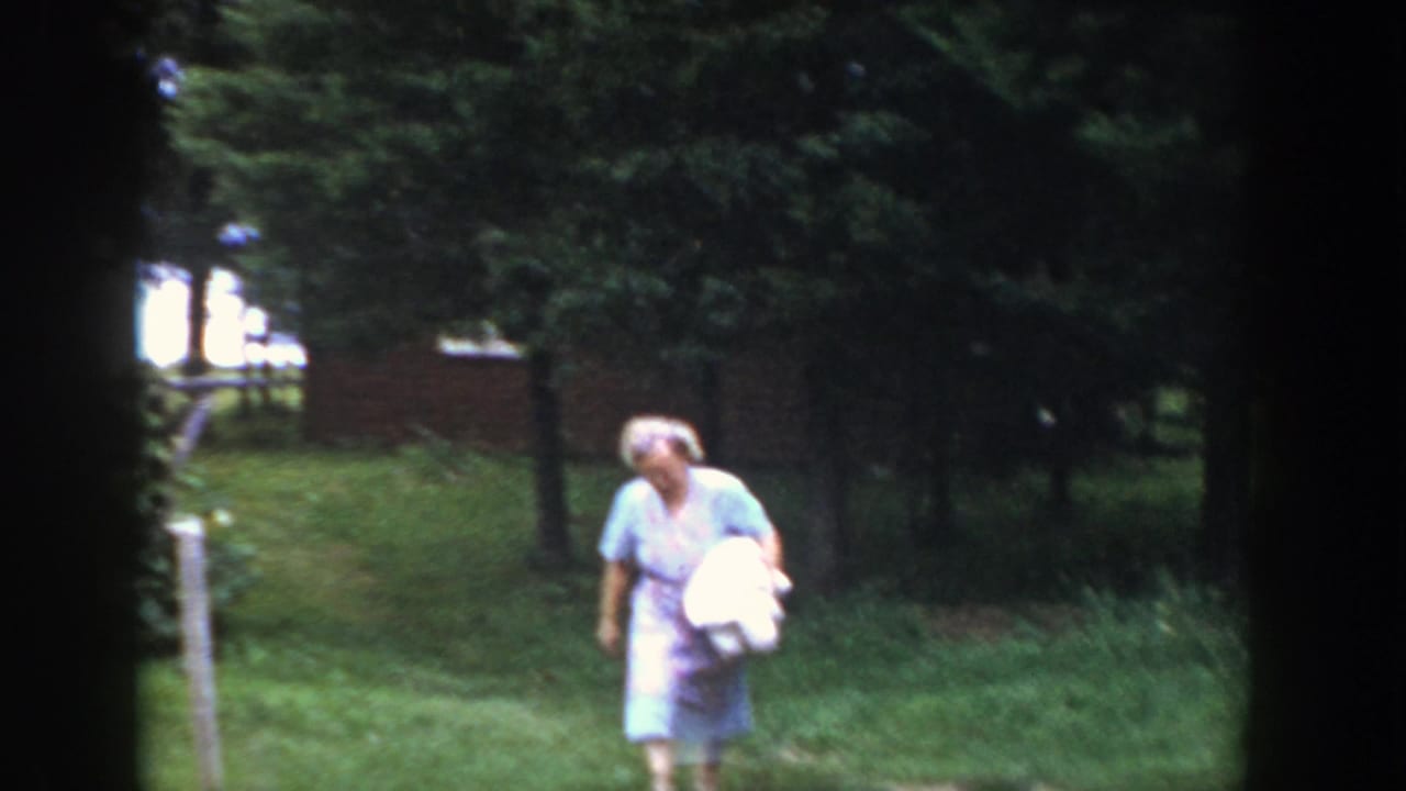 Woman in a light dress carries a white bundle across a grassy lawn near trees.