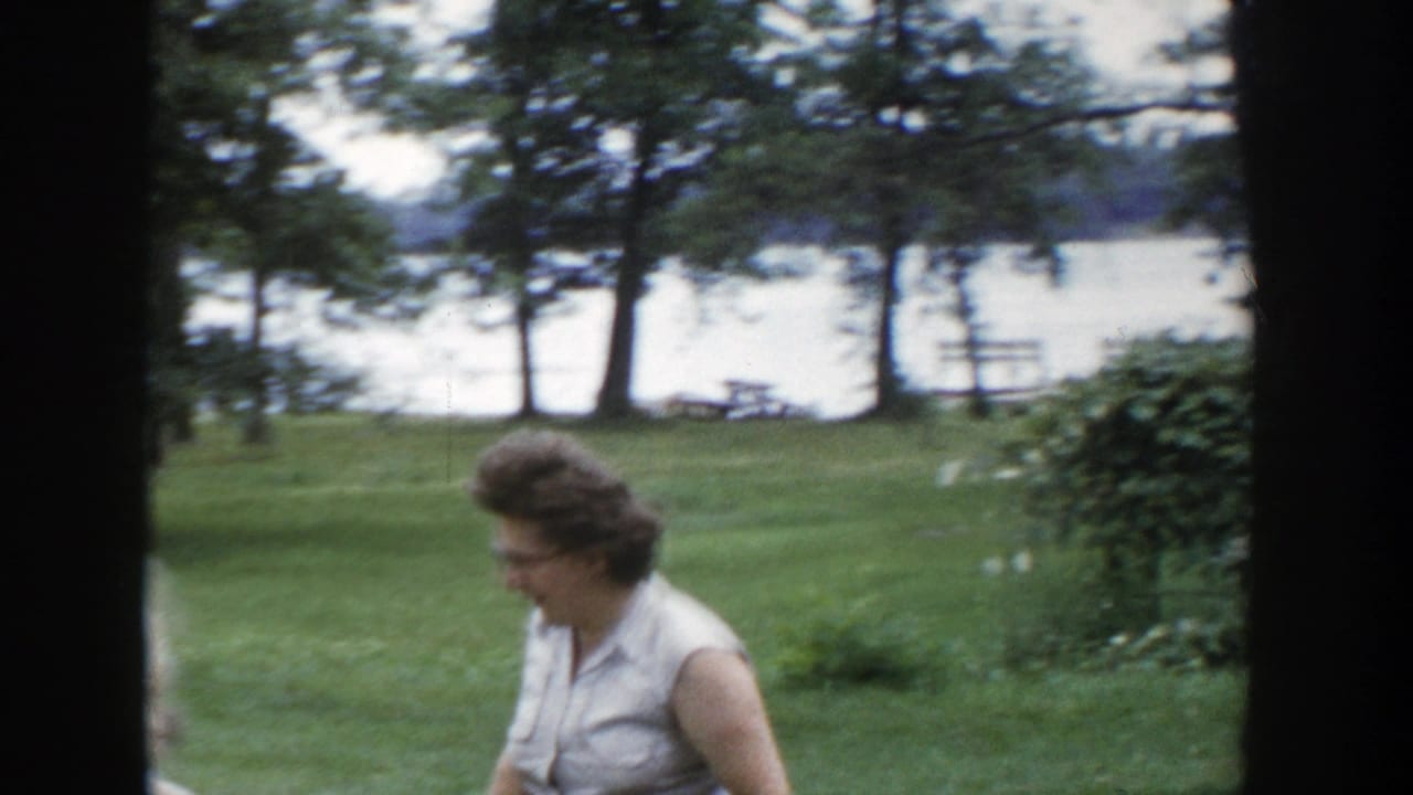 Woman in a sleeveless light dress walks across a lawn with lake and trees behind her.