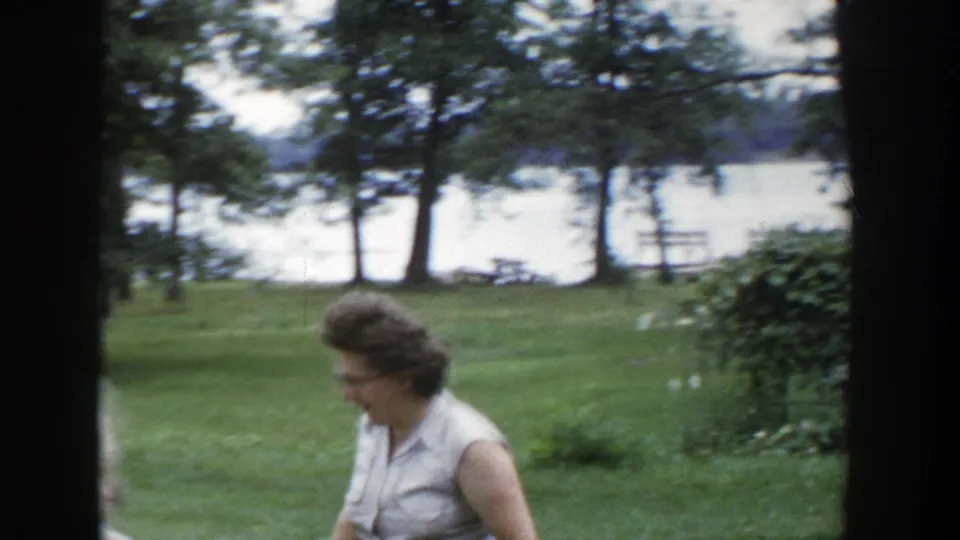 Woman in a light sleeveless dress moves through a Wisconsin lakeside lawn scene in 1953 home-movie footage.