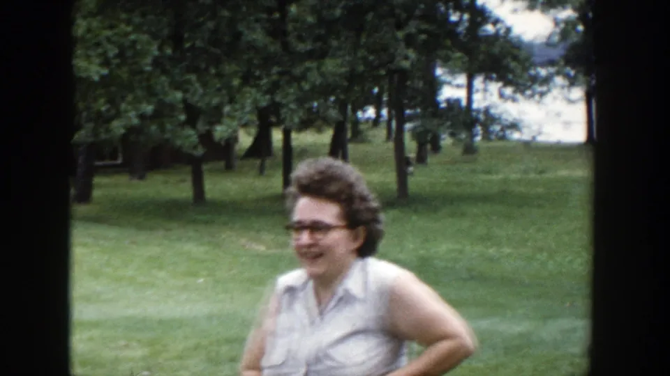 Smiling woman with glasses appears on a tree-lined lawn beside a Wisconsin lake in 1953 8mm footage.