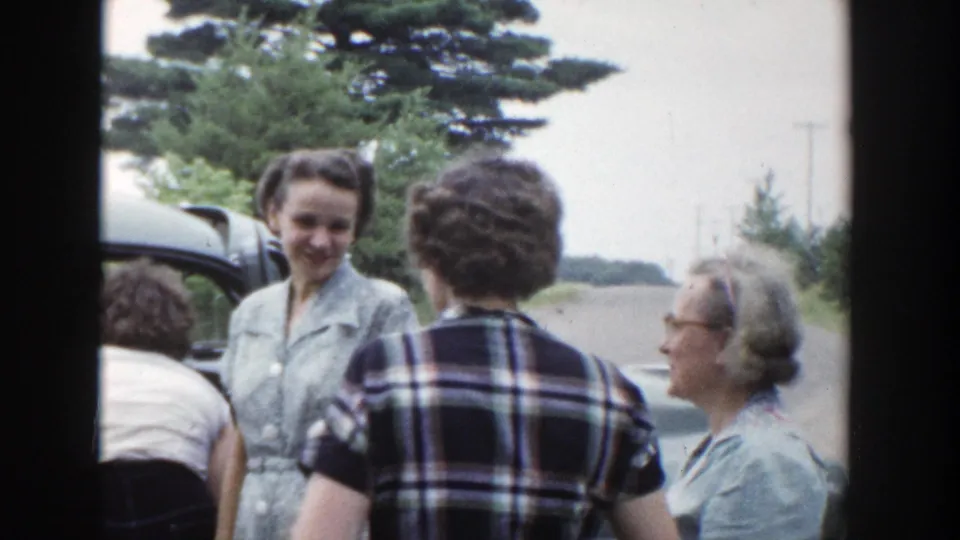 Group of women in mid-century dresses gathers near a car by a Wisconsin lakeside road in 1953 home-movie footage.