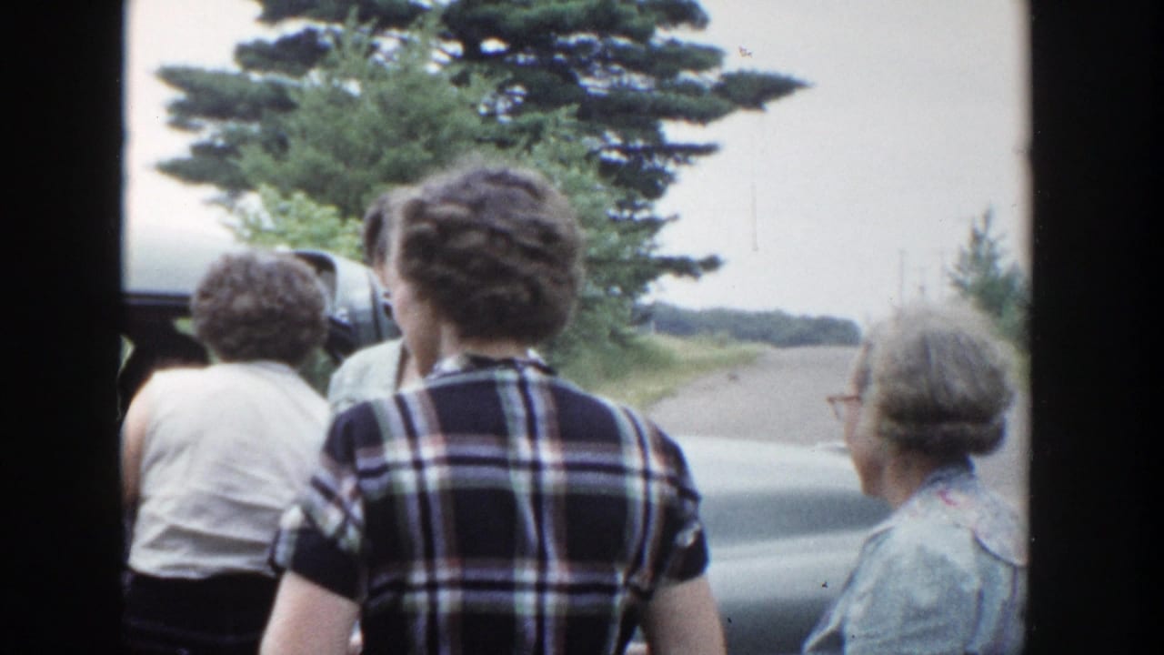 1953 Wisconsin home-movie frame showing women from behind near a car on a tree-lined rural road.