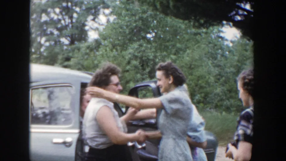 Two women appear to embrace beside a car in a rural Wisconsin farewell, with others partly visible nearby.