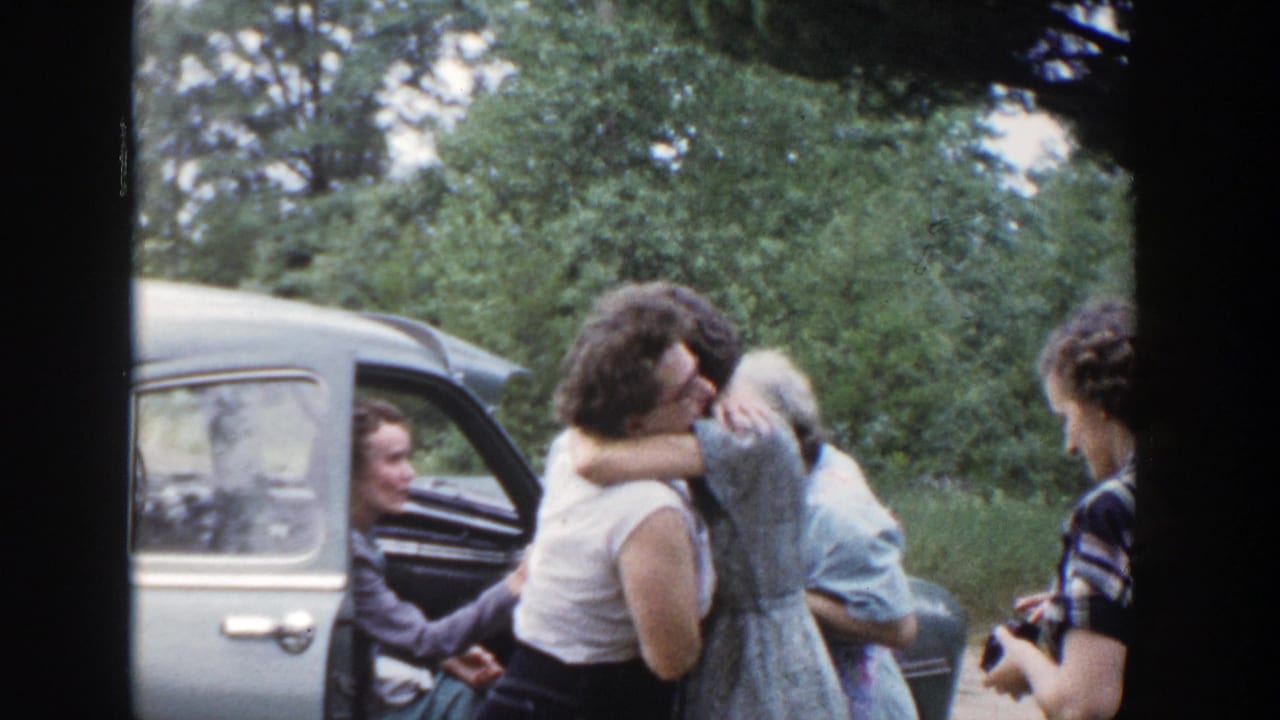 In Wisconsin 1953 home-movie footage, adults greet with a hug beside a light-colored car parked near trees.