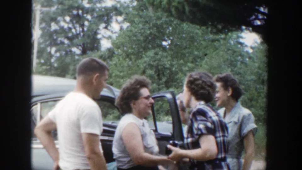 A man and several women talk beside a parked car during a 1953 Wisconsin outdoor greeting scene.