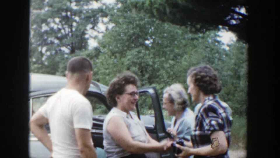 Adults smile and converse near an open car door in 1953 Wisconsin home-movie footage, with trees in the background.