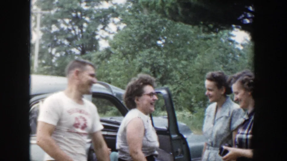 The group continues laughing beside the car, with the SHAWANO shirt text and open vehicle door visible in the Wisconsin home-movie frame.