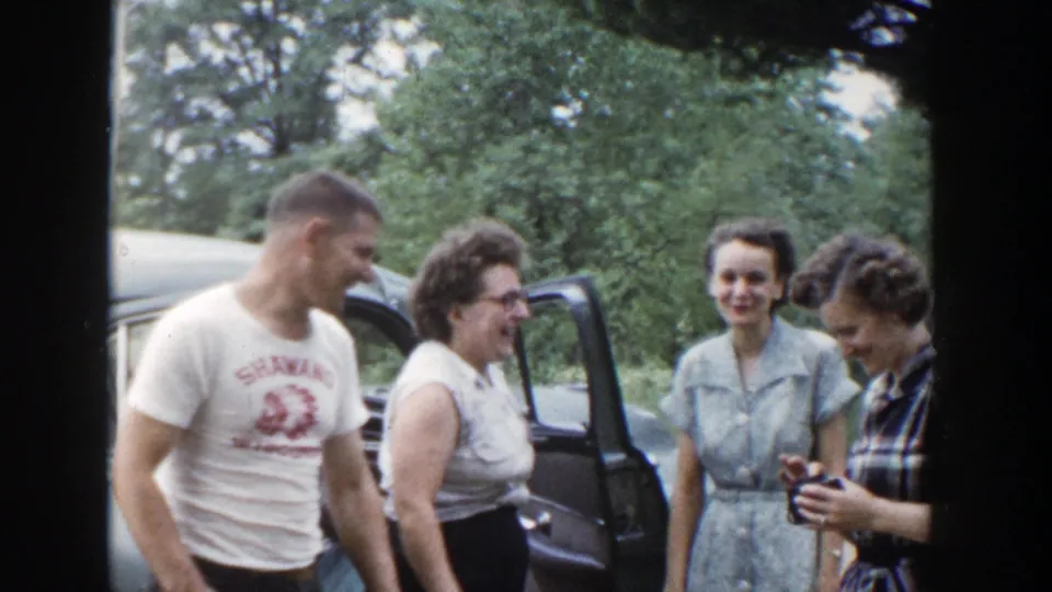 A man and three women stand beside the car as one woman looks down at a small object, continuing the casual 1953 Wisconsin gathering.