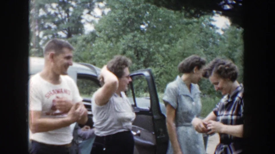 Final frame shows the adults beside the parked car, with one woman raising an arm and others looking down during the informal outdoor exchange.