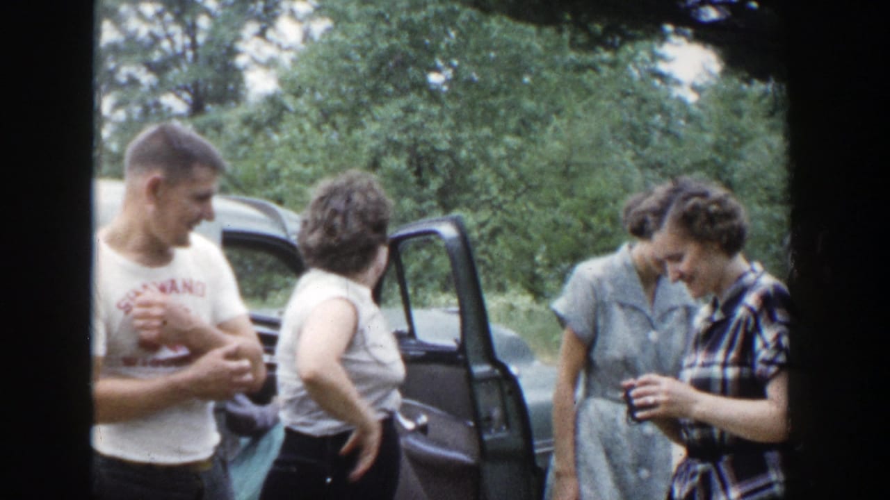 Four adults stand beside a blue car with an open door in a wooded outdoor area.