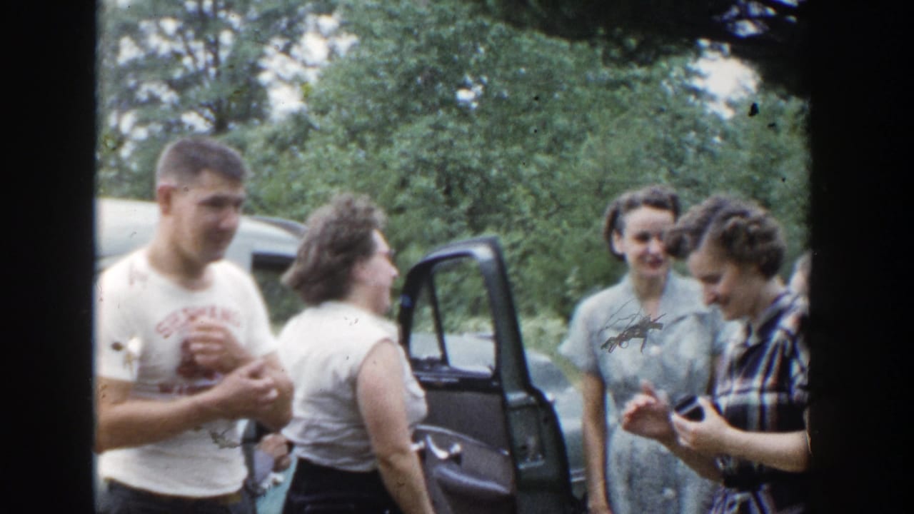Adults talk and smile beside an open car door with trees in the background.