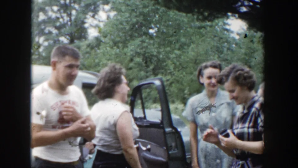 Adults in casual 1950s clothing appear to chat near a blue car in a wooded Wisconsin setting.