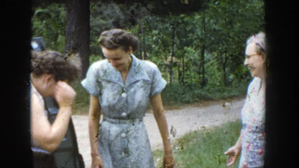 Women laugh and talk near the parked car in a wooded Wisconsin roadside scene from 1953 home-movie footage.