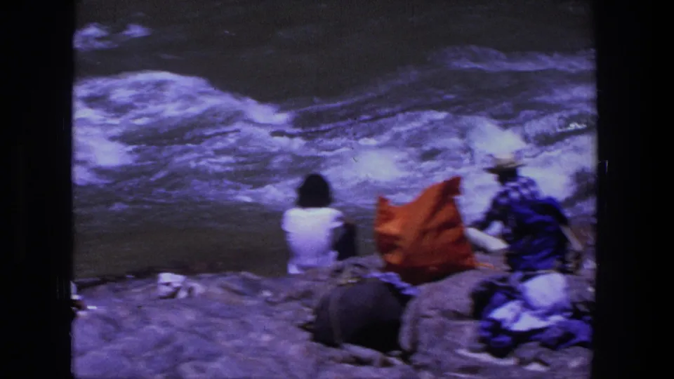 1969 Utah riverbank view with seated people, an orange bag, and a man in a blue shirt near the current.