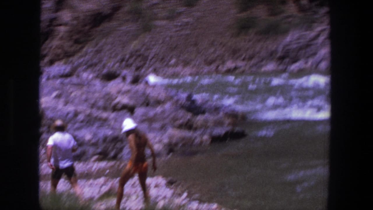 Two adults wade near a rocky riverbank with shallow water behind them.