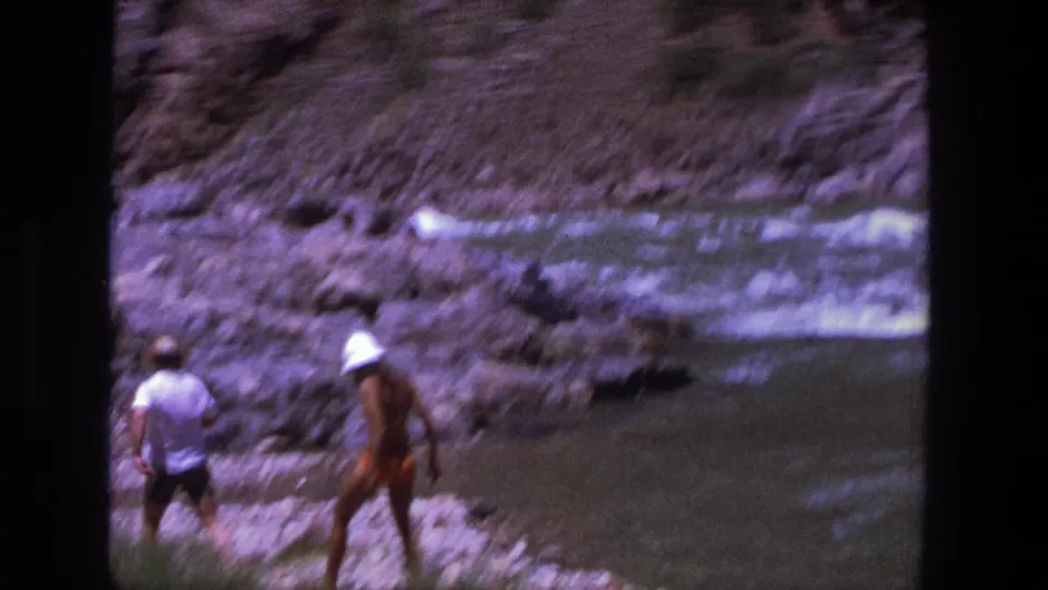 1969 Utah 8mm frame showing two adults near a rocky riverbank, one in a white shirt and another in bright swimwear by shallow water.