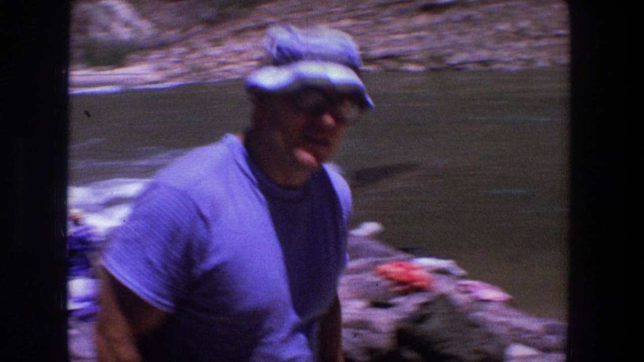 Man in a blue shirt and brimmed hat stands near a riverbank.