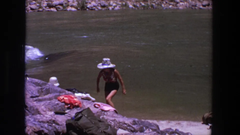 1969 Utah 8mm frame showing a woman in a dark swimsuit and white hat stepping across rocks near personal belongings and river water.