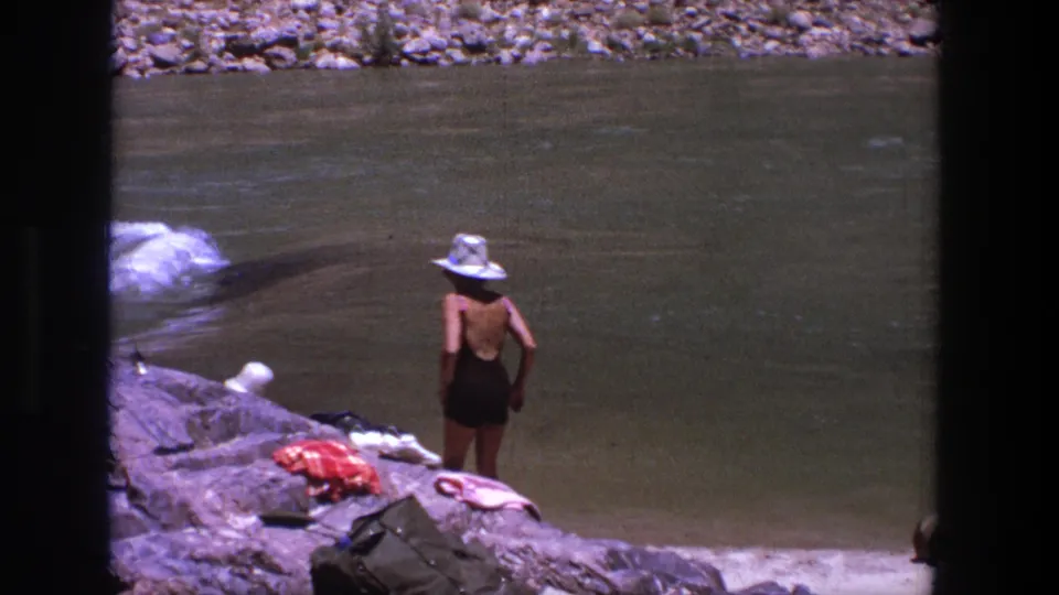 1969 Utah home-movie frame with a woman in a dark swimsuit and white hat facing the river beside clothing and bags on the rocks.