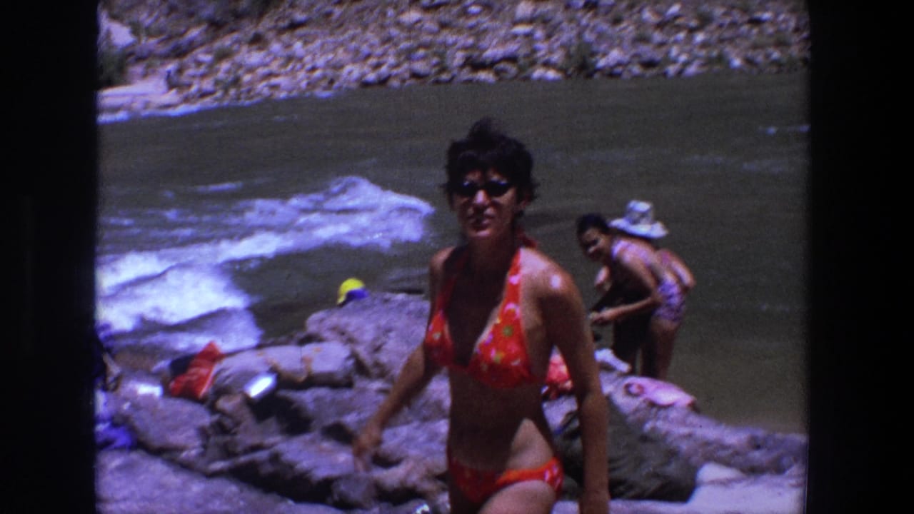 Woman in a red floral bikini stands on rocks near a rushing river while another adult is behind her.