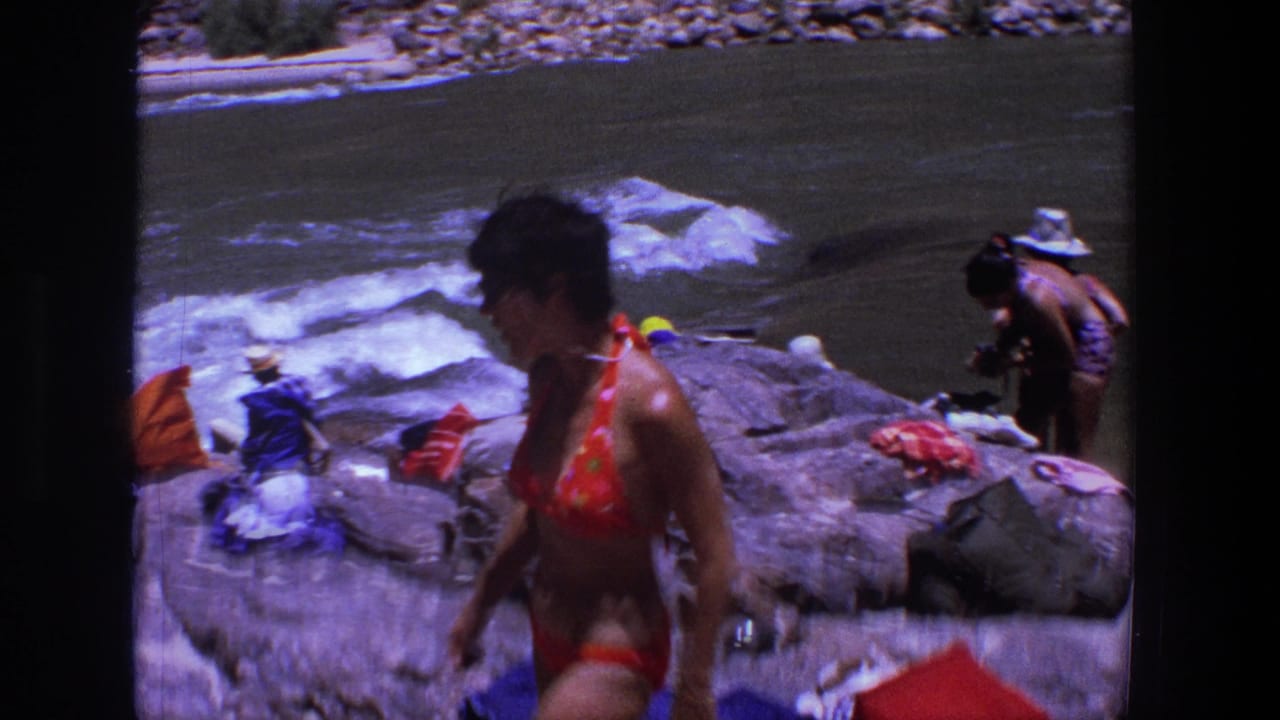 Woman in a red bikini walks across river rocks with towels and people near the water behind her.