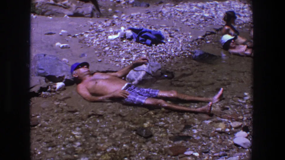 A man in a blue cap and plaid shorts lies in shallow water on a rocky Utah riverbank, with another adult sitting nearby.