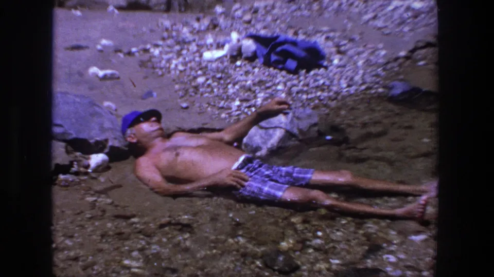 In the closing frame, a man reclines in shallow river water on a rocky Utah shore while towels and belongings sit nearby.