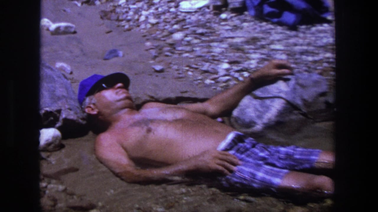 Shirtless man in a blue cap lies on rocky wet ground beside scattered stones.