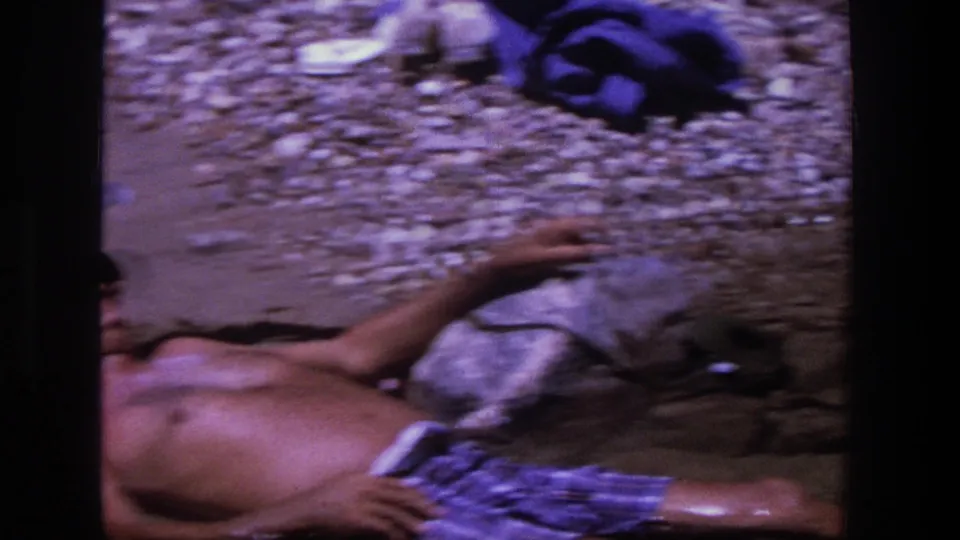 A handheld 8mm frame crops the reclining man at the shoreline, emphasizing wet rocks, pebbles, and plaid swim trunks.