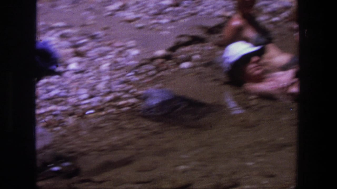 Adults relax in shallow river water at a rocky Utah bank in 1969, with hats and swimsuits visible in soft 8mm color.