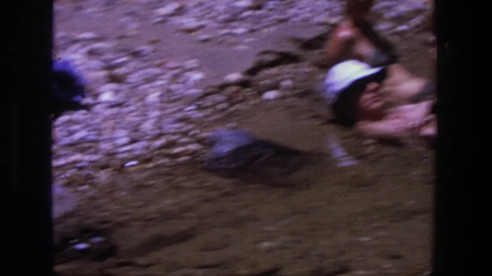 Adults relax in shallow river water at a rocky Utah bank in 1969, with hats and swimsuits visible in soft 8mm color.