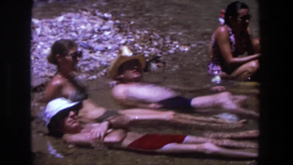 Several adults in swimsuits, hats, and sunglasses relax in shallow Utah river water in this 1969 home-movie frame.
