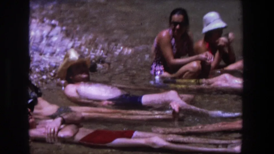Adults in vintage swimwear rest and talk in shallow river water, with stones and sun hats visible in the Utah 1969 footage.